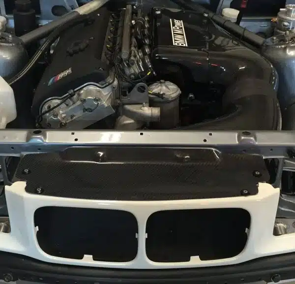 Car engine bay with a visible engine and black intake parts, viewed from the front grill area.