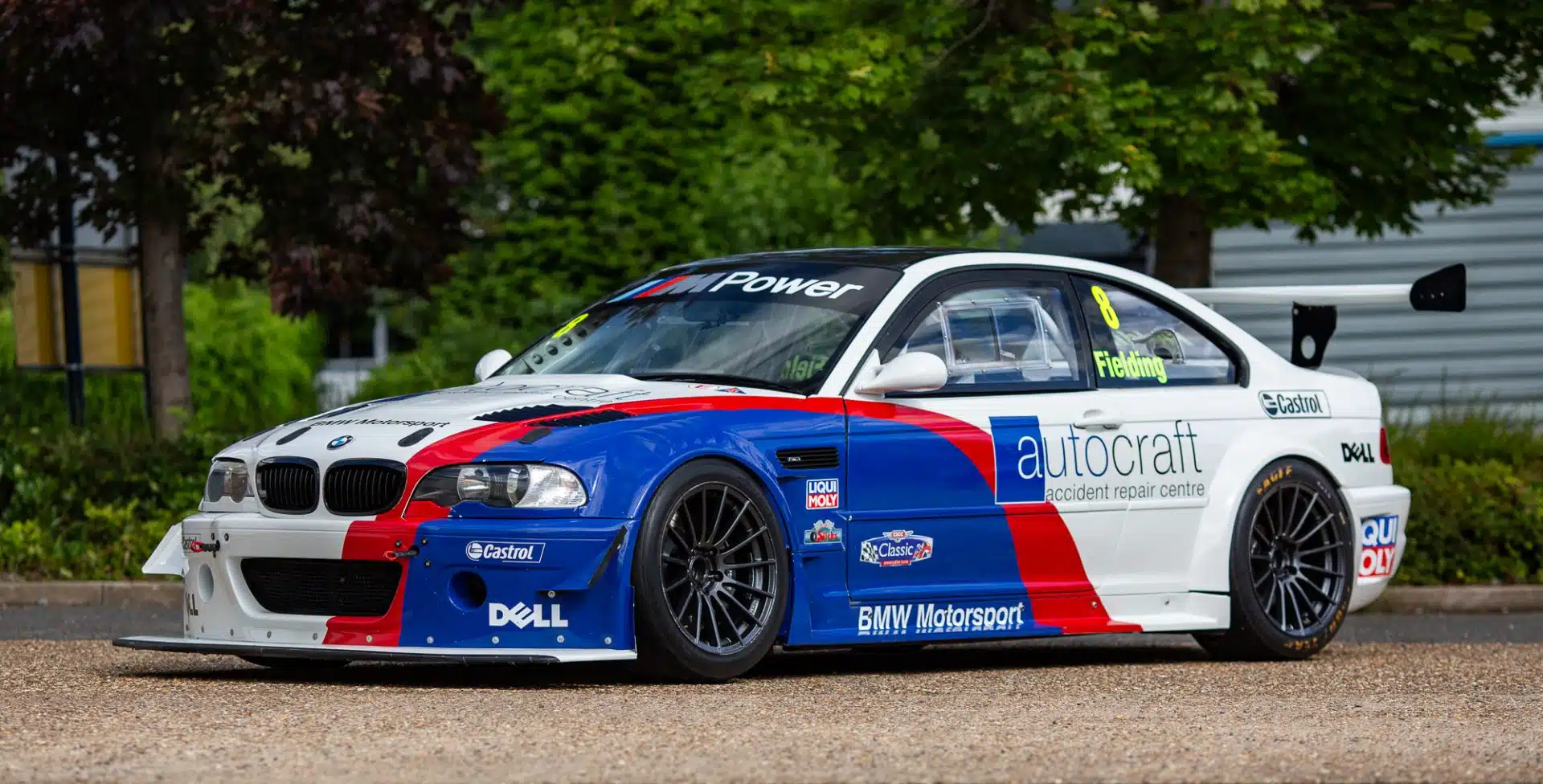 White BMW E46 M3 race car with blue and red graphics parked outdoors.