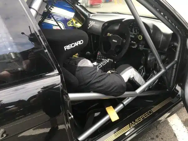 Race car driver in full gear sitting inside a car with a roll cage and Recaro seat.
